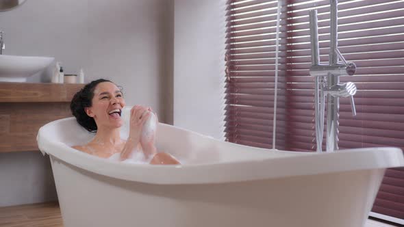 Young Funny Dynamic Emotional Woman Lies in Foam Bath Enjoys Music Sings Song Using Shampoo Bottle