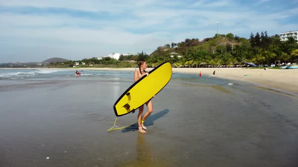 Young woman surfer walking on tropical ocean beach holding yellow surfboard, attractive caucasian mo