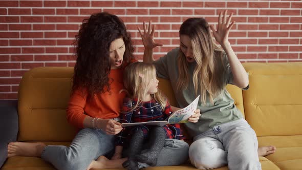 Two Women Read a Book Together to a Small Preschool Girl Sitting on the Sofa at Home