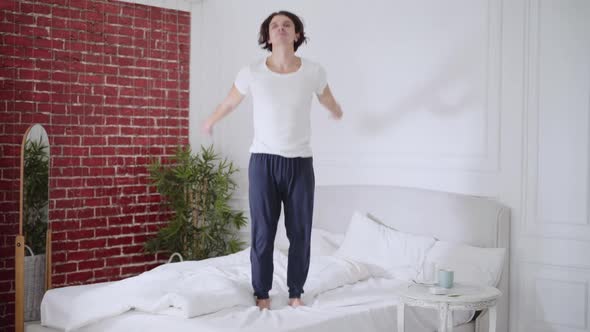 Wide Shot of Happy Carefree Young Man Jumping on Bed Smiling