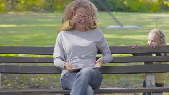 Young Pretty Caucasian Woman Sitting on the Bench with a Tablet, Two Little Cute Girls Coming