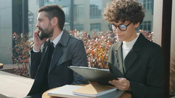 Ambitious Businessman Talking on Mobile Phone and Consulting Female Colleague Sitting in Street Cafe