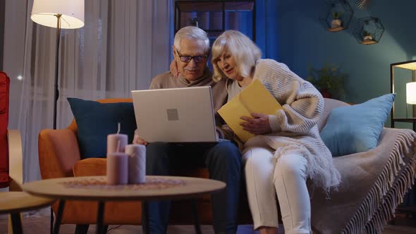 Senior Grandparents Couple Reading Book Using Laptop Pc on Couch in Night Living Room at Home