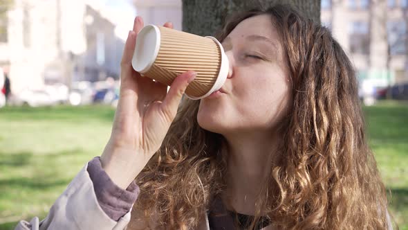 Young Woman Drinks Coffee in Paper Cup Sitting in City Park