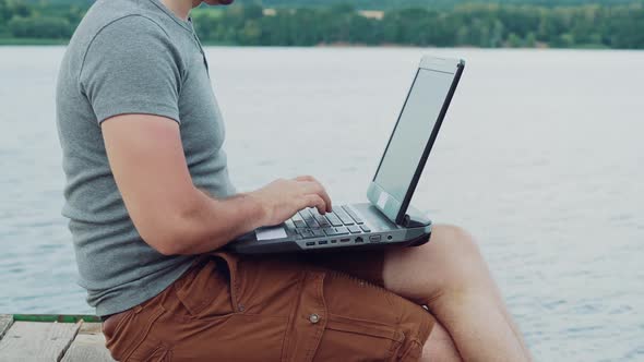 Handsome man typing on a laptop sitting over the flowing river and enjoys the nature in summer