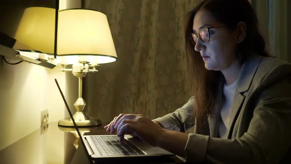 Young Woman in Glasses and a Suit is Typing Text on a Laptop Under the Light of a Table Lamp