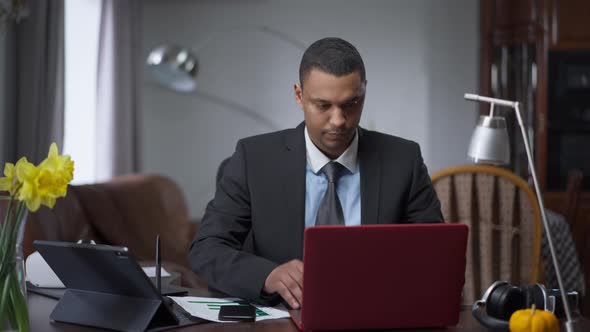 Confident Young African American Man Opening Laptop and Typing on Keyboard Sitting in Living Room at