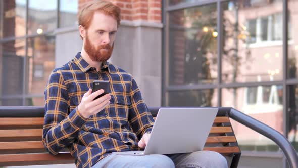 Redhead Beard Young Man Using Smartphone and Laptop Sitting on Bench