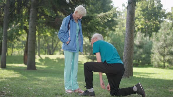 Thankful Senior Wife Hugging Husband Tying Shoelace on Sneakers in Spring Summer Park