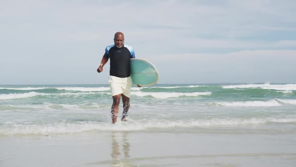 African american senior man walking on a beach holding surfboard and running out of the sea