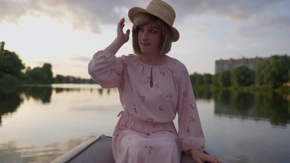 Charming Caucasian Trans Woman in Pink Dress and Straw Hat Sitting in Boat on Lake Looking Away