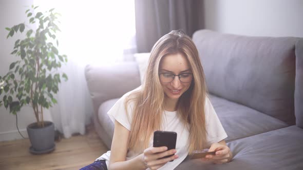 Blonde Woman Sitting on the Floor Pays for Purchases Online with a Credit Card and Phone Home