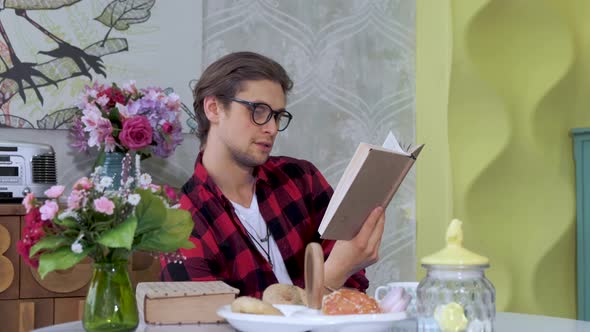 Portrait of young caucasian man sitting at table in modern spacious kitchen, reading book