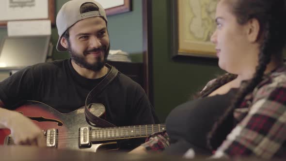 Young Smiling Bearded Man Playing Guitar in the Bar, Attractive Plump Woman Sitting Near Singing