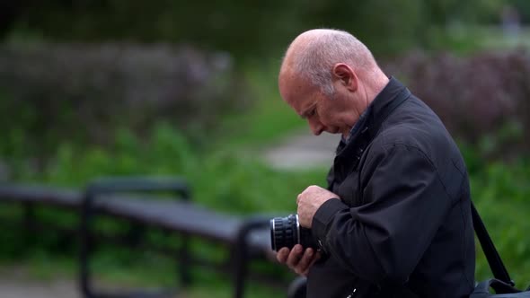 An Elderly Bald Man in Nature, Blurred Green Background of the Park and Forest, He Holds a Camera in