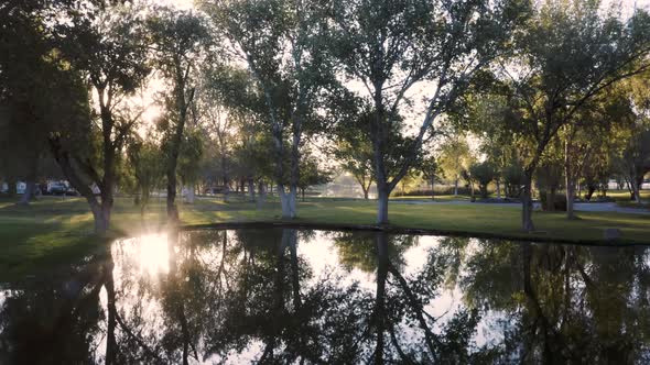 Flying over beautiful park pond with reflection of trees toward setting sun