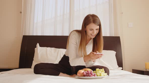 Happy Young Woman Sitting on Bed with Plate of Fresh Fruits and Talking on