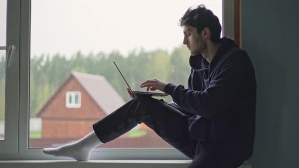 A Young Man is Typing on a Laptop Sitting at the Window Against the Backdrop of a Picturesque Rural