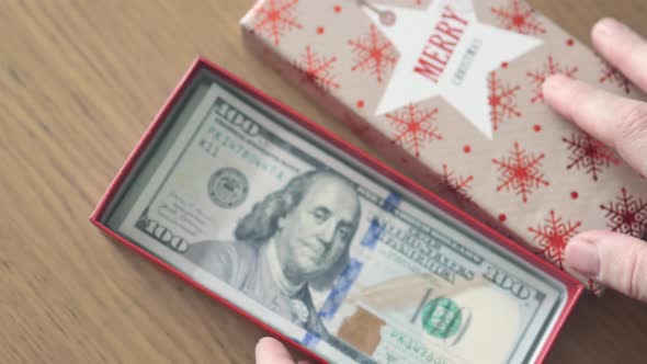 Hand Putting Hundred US Dollar Banknote Bill in Present Gift Box with Words Merry Christmas on It