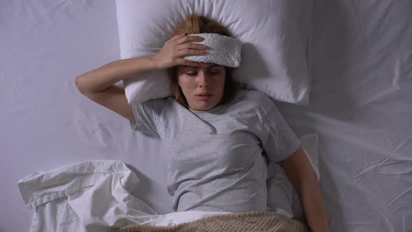 Upset Woman With Compress Lying in Bed, Suffering From Migraine Taking Pill