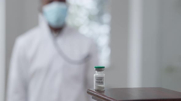 Covid19 Vaccine Jab on Table with Blurred African American Doctor Walking From Background Taking