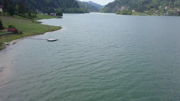 Aerial Panning Shot of edge of lake with a cabin and trees in the background and a pier in the foreg