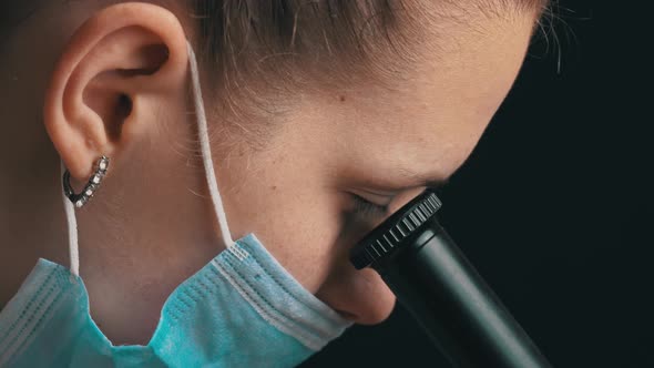 Girl Scientist Looks Into the Lens of a Microscope in a Mask Closeup