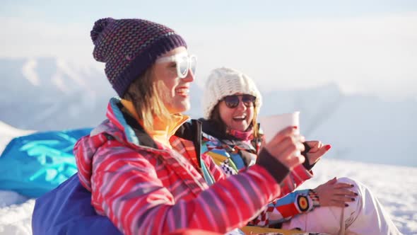 Two Caucasian Females Wearing Glasses and Knitted Hats Sitting at Ski Mountain Resort Talking