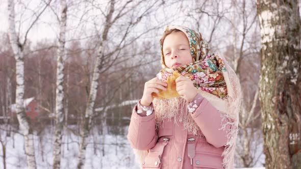 Cute girl in a traditional Russian headscarf with bagels eats pancake on winter background.