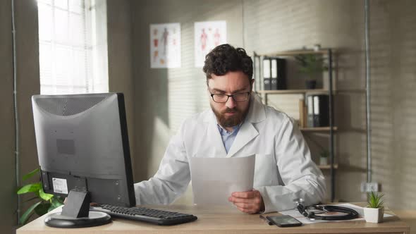 Experienced Caucasian Adult Doctor with Beard are Sitting at Desk and Studying Patients Reports