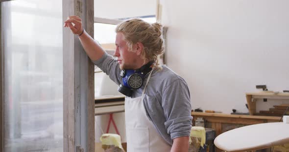 Caucasian male surfboard maker wearing a face mask and standing in his studio