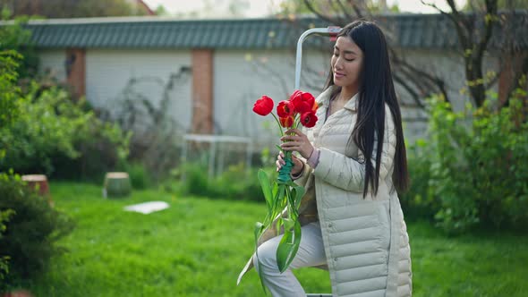 Wide Shot of Happy Young Asian Female Florist Standing in Sunny Garden with Bouquet of Red Tulips