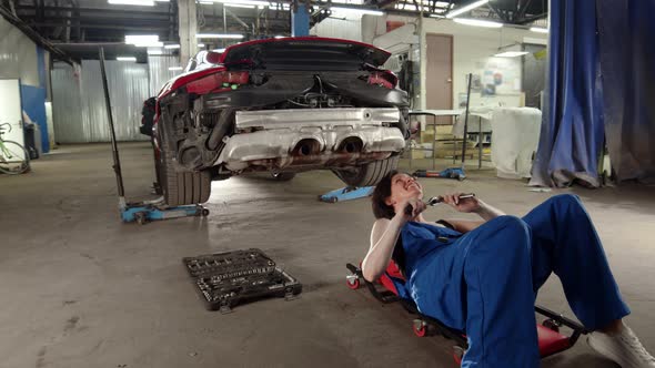 Woman Mechanic in Car Service Rolls Under Sports Car with Wrench or Ratchet in Hands and Repairs Car