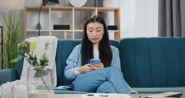 Asian Girl with Black Hair in Casual Clothes Sitting on Soft Couch at Home and Texting on Mobile