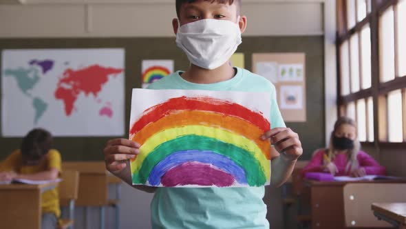 Boy wearing face mask holding a rainbow painting in class at school 