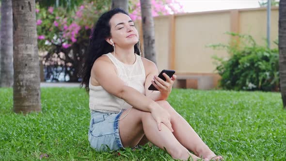 European Woman Use By Mobile Phone Listen Music Via Headphones on Nature Relax in the Park Sitting