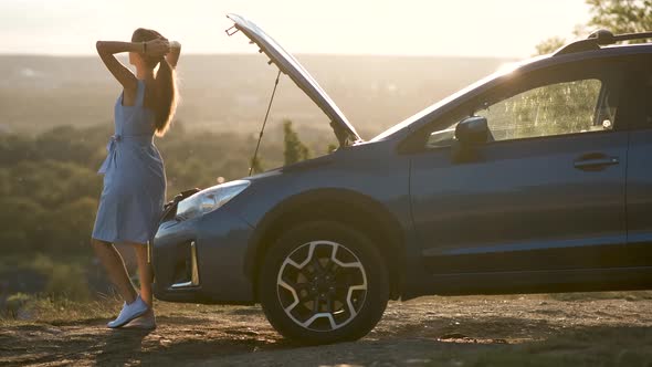 Young female driver standing near a broken car with open up hood inspecting her vehicle engine 