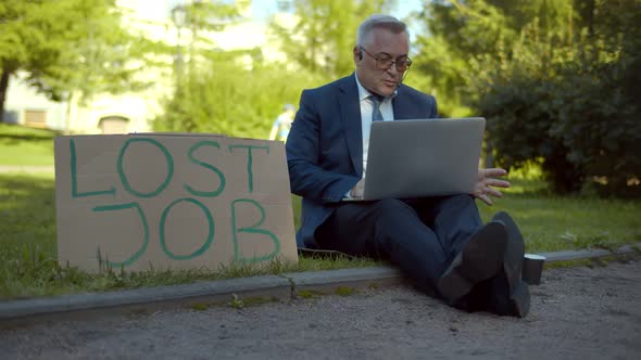 Aged Businessman Having Video Call on Laptop Sitting on Grass with Lost Job Sign and Cup for Charity