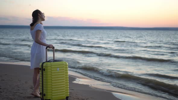 Young Woman with a Yellow Suitcase on the Beach By the Sea. A Girl in a White Dress Looks at the