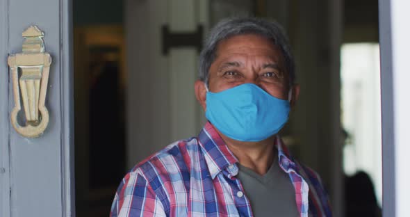 Senior mixed race man wearing face mask at open front door greeting