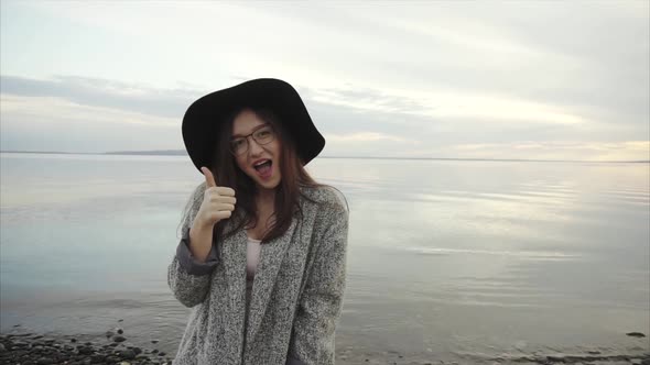 Young Pretty Woman in Black Hat and Glasses Showing Thumb Up Near Sea at Sunset