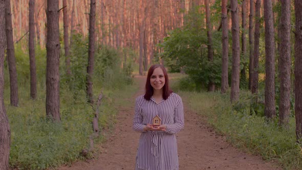 Young Woman with Small Wooden House in Hands Stands in Forest