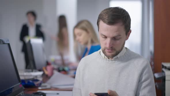 Brunette Caucasian Man Sneezing Surfing Internet on Smartphone in Office and Blurred People Leaving
