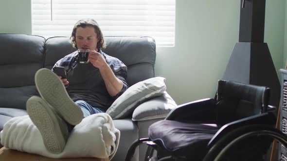 Happy caucasian disabled man sitting on sofa in living room using smartphone and drinking coffee