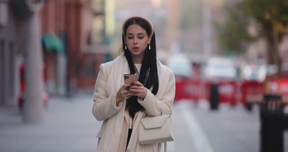 Young Stylish Woman Looks Into Smartphone Walking in City