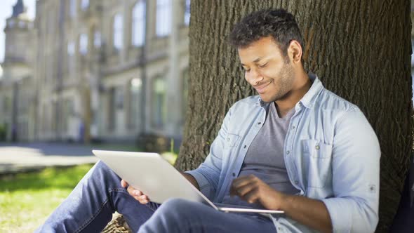 Multiracial College Student Sitting Under Tree With Laptop, Checking Final Paper