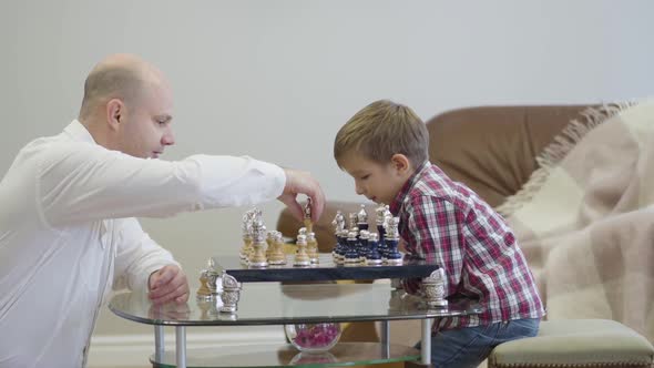 Side View of Caucasian Man in White Shirt Sitting in Front of Chess Board and Playing with Clever