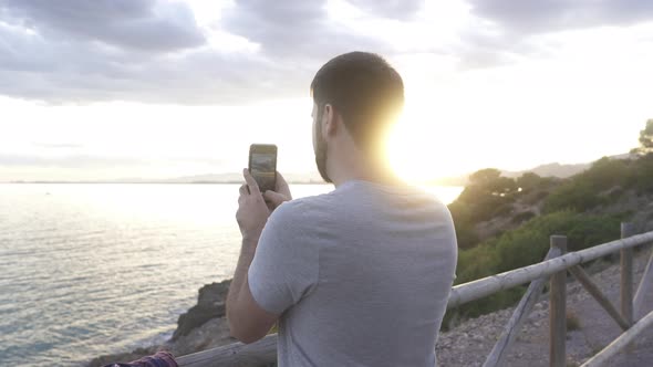 A man takes pictures of the sunset with the cell phone on the coast, the sea and the golden sky are