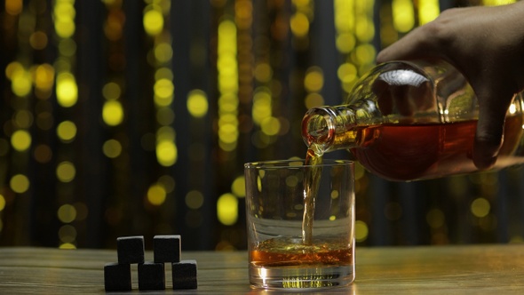 Pouring of Golden Whiskey, Cognac or Brandy From Bottle Into Glass with Rocks Ice Stones on Table