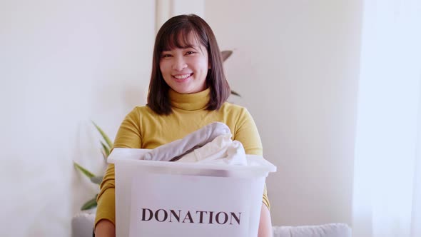 Portrait of young Asian woman looking at camera holding box with clothes for donation. Concept campa